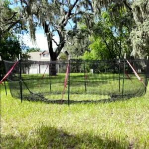 9:16 vertical medium-close view of The P.I.G. Hog Trap™ at ground level with a home in the background, showcasing a complete sounder removal system designed to help protect homes, property, and surrounding land from feral hog damage.