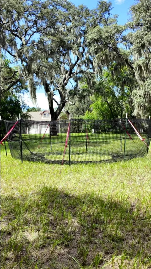 9:16 vertical medium-close view of The P.I.G. Hog Trap™ at ground level with a home in the background, showcasing a complete sounder removal system designed to help protect homes, property, and surrounding land from feral hog damage.